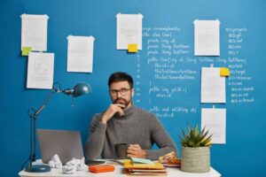 A person sits at a desk with a laptop and documents, surrounded by notes and a potted plant against a bright blue wall.