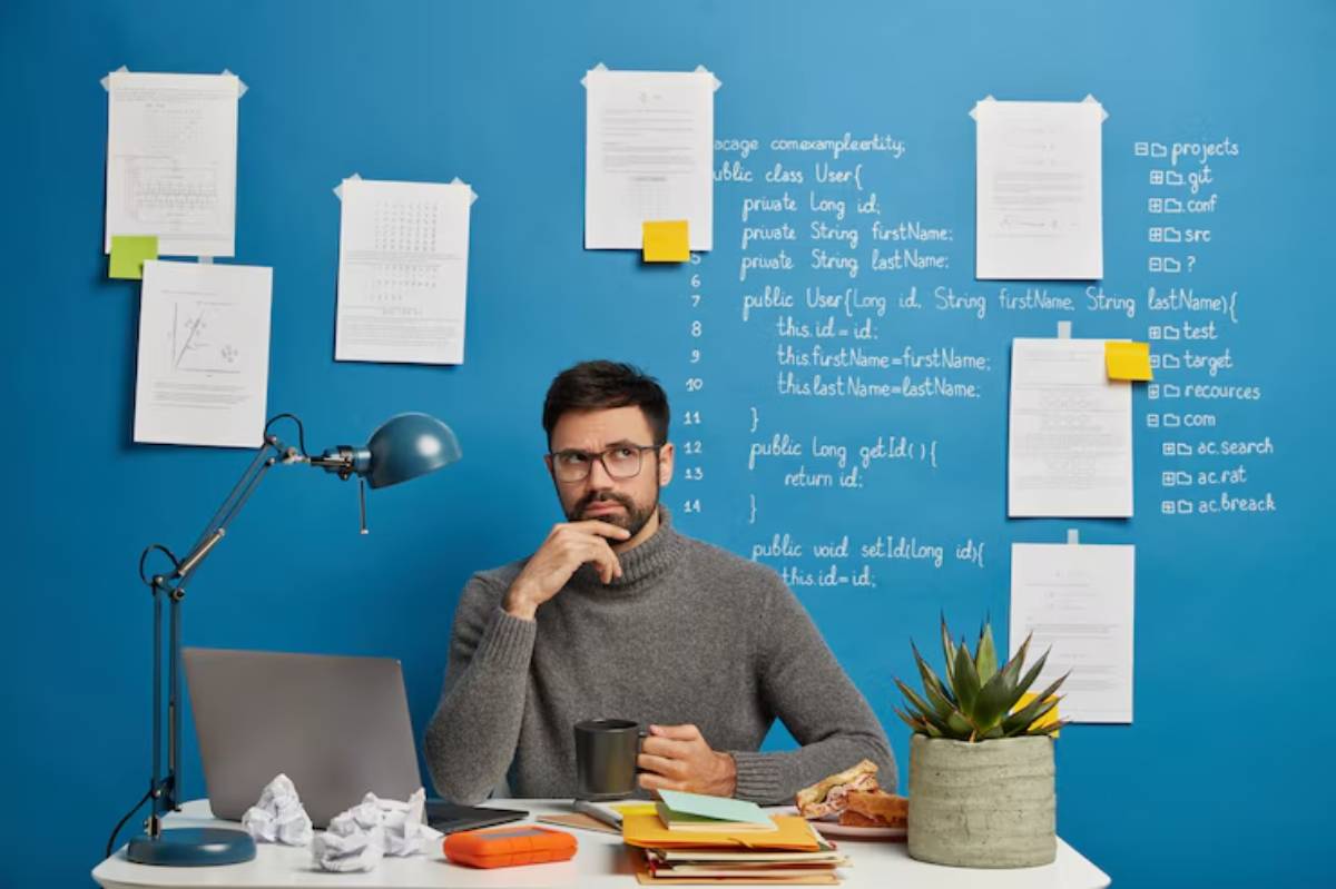 A person sits at a desk with a laptop and documents, surrounded by notes and a potted plant against a bright blue wall.