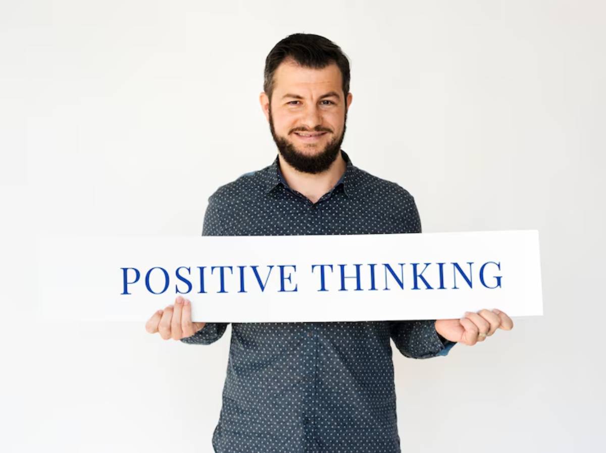 A person in a polka-dotted shirt holds a sign that reads POSITIVE THINKING against a plain white background.
