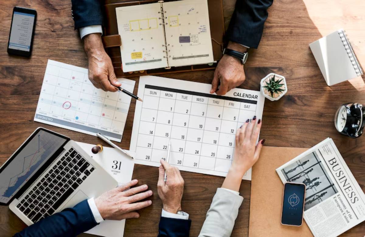 A top-down view of a meeting with hands pointing at a calendar, a laptop, and various business documents on a wooden table.