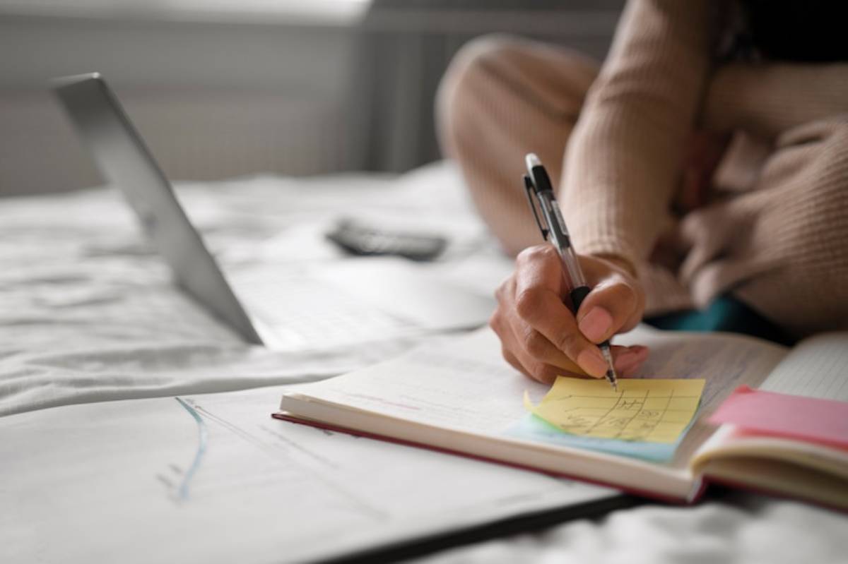 A person writes on a sticky note placed on a notebook while seated on a bed, with a laptop and calculator nearby.