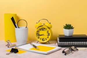 A vibrant workspace with a yellow clock, white planter, notebooks, pens, and glasses, all against a cheerful yellow background.