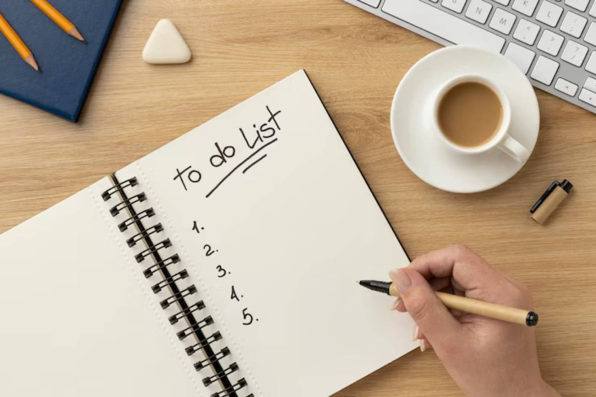 A hand writes on a blank to-do list in a notebook beside a coffee cup, pencils, and a keyboard on a wooden desk.