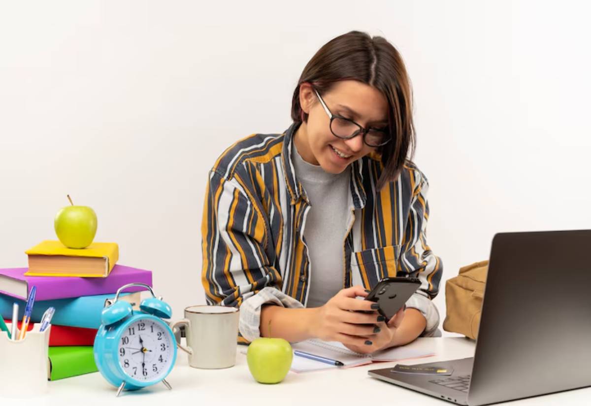 A student sits at a desk with a laptop, books, a cup, an alarm clock, and an apple, checking their phone thoughtfully.