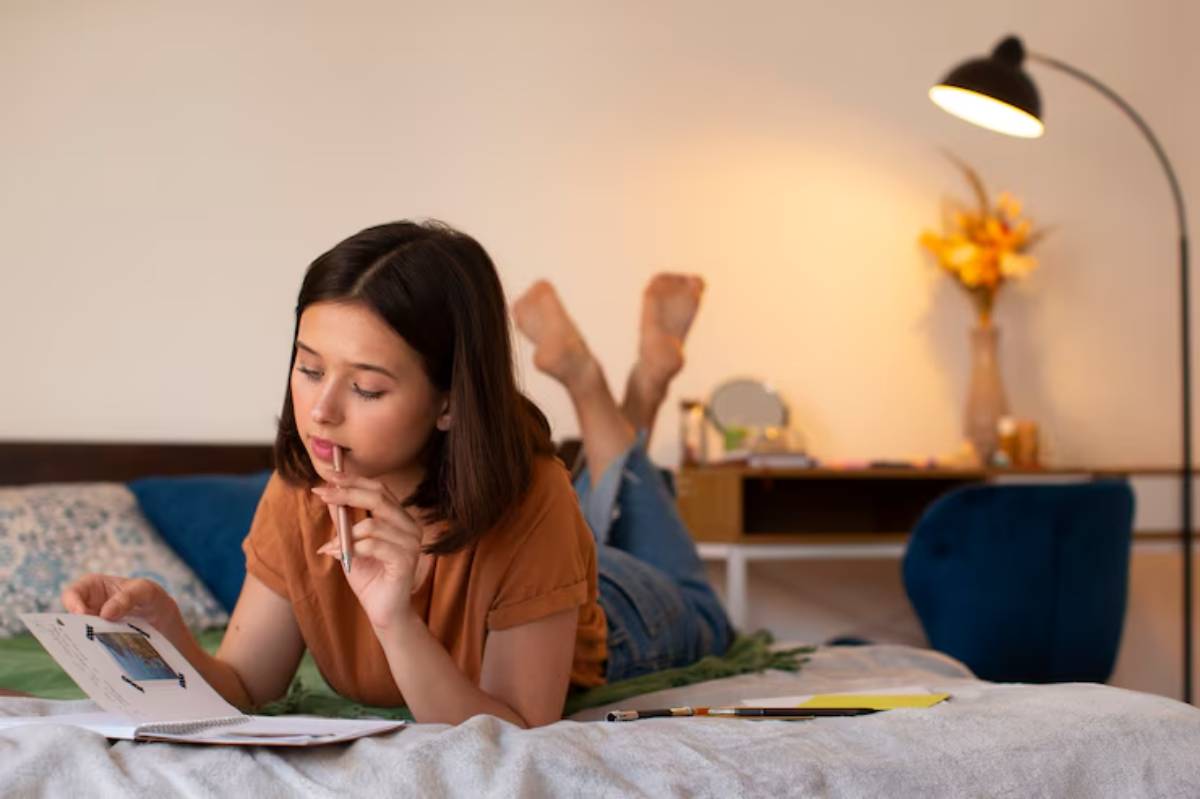 A person lies on a bed, reading and taking notes, surrounded by a cozy, well-decorated room with a warm lamp and flowers.
