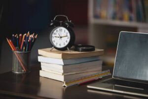 A black alarm clock sits atop stacked books next to a laptop, colorful pencils in a cup, and a ruler on a wooden desk.