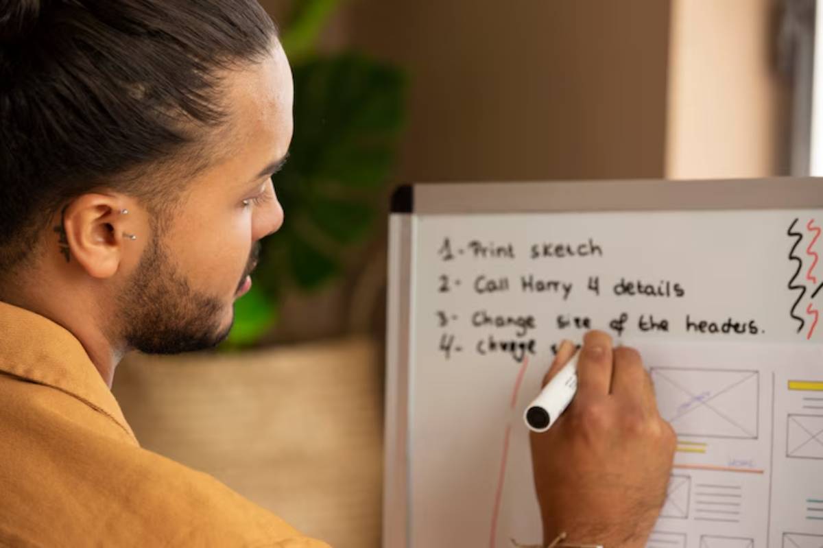 A person wearing a brown shirt writes notes on a whiteboard with design instructions and a checklist in a well-lit room.