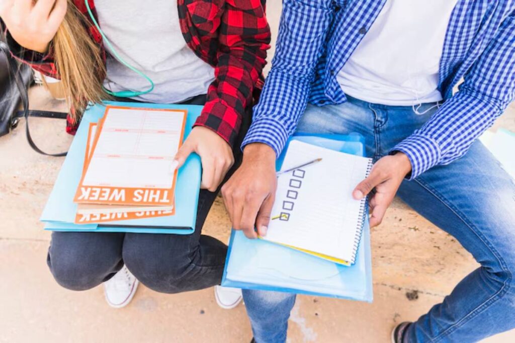 Two students sitting side by side, reviewing planners and notes, with casual attire and headphones on, engaged in study.