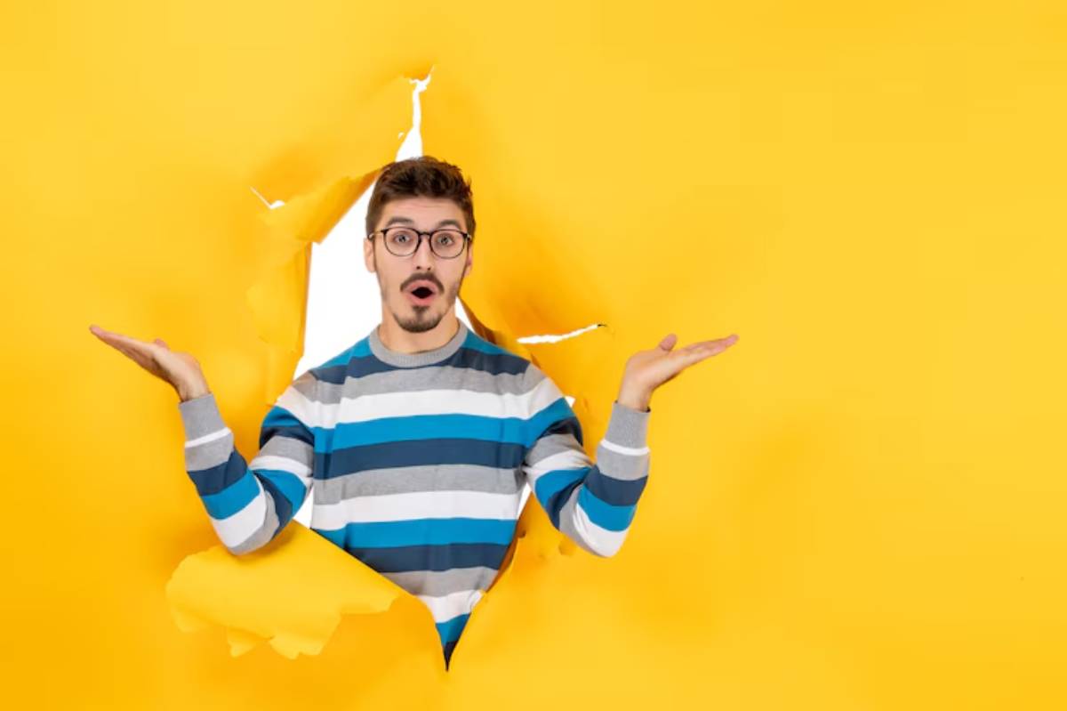 A person in a striped sweater emerges through a torn section of a bright yellow wall, arms outstretched in a playful gesture.