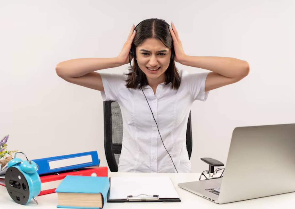 A woman sits at a desk, wearing headphones and holding her head with both hands, surrounded by books and office supplies.