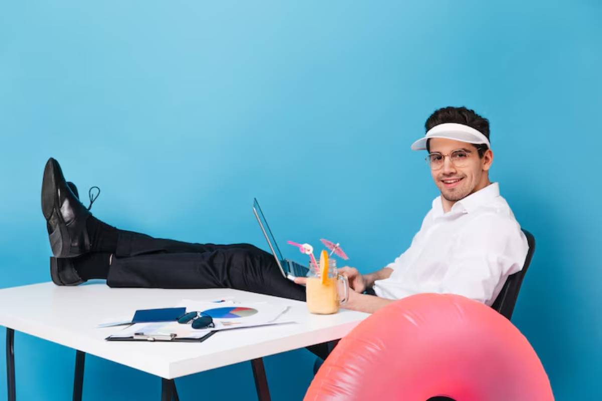 A person in a white shirt relaxing at a desk, with feet up, laptop open, and a colorful drink beside a pink inflatable ring.