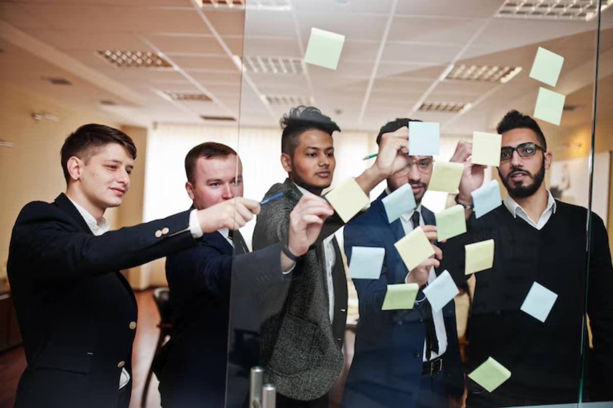 A group of professionals in business attire actively placing colorful sticky notes on a glass wall inside an office environment.