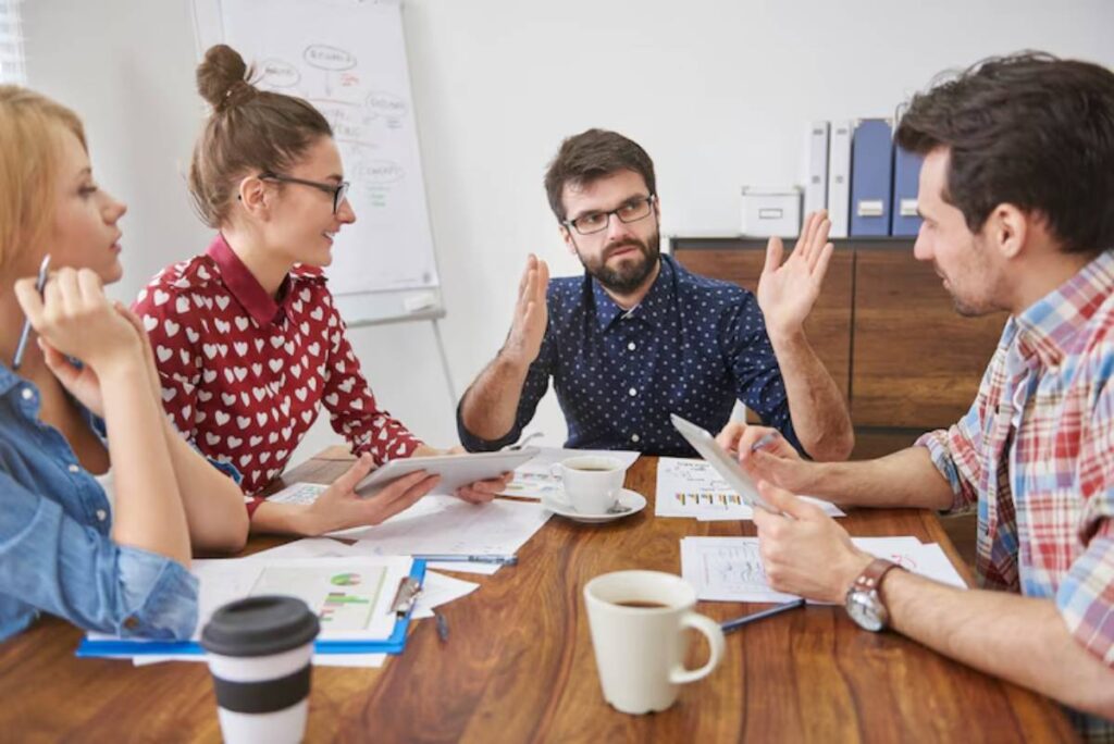 A group of professionals engaged in a discussion around a wooden table, with papers, a tablet, and coffee cups present.