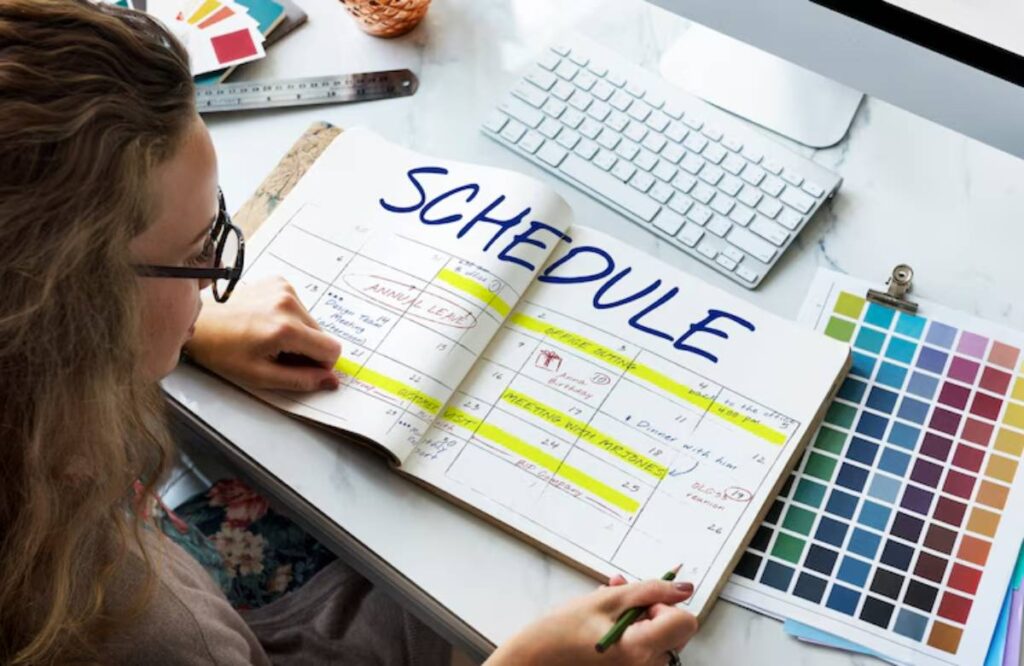 A hand holds a pen over an open planner labeled "SCHEDULE," filled with colorful highlights and notes, beside a computer keyboard and color palettes.
