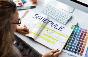 A hand holds a pen over an open planner labeled "SCHEDULE," filled with colorful highlights and notes, beside a computer keyboard and color palettes.