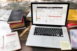 A laptop displaying a calendar sits on a wooden desk surrounded by books, notebooks, a ruler, and stationery.