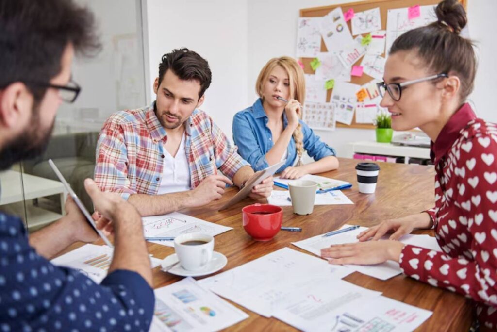 A group of professionals engaged in a meeting, discussing documents and data on a wooden table, with coffee cups and notes present.