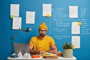 A person wearing a yellow beanie sits at a desk with a laptop, surrounded by notes and coding examples pinned to a blue wall.