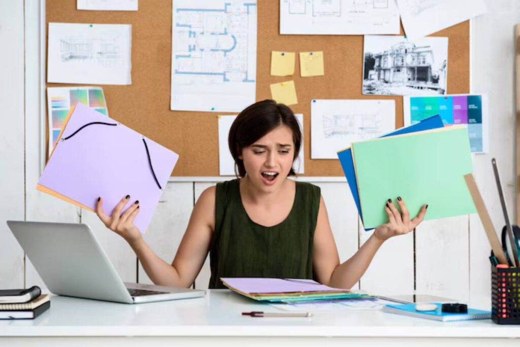 A person sits at a desk, holding colorful folders in each hand, surrounded by a laptop and architectural plans on a bulletin board.