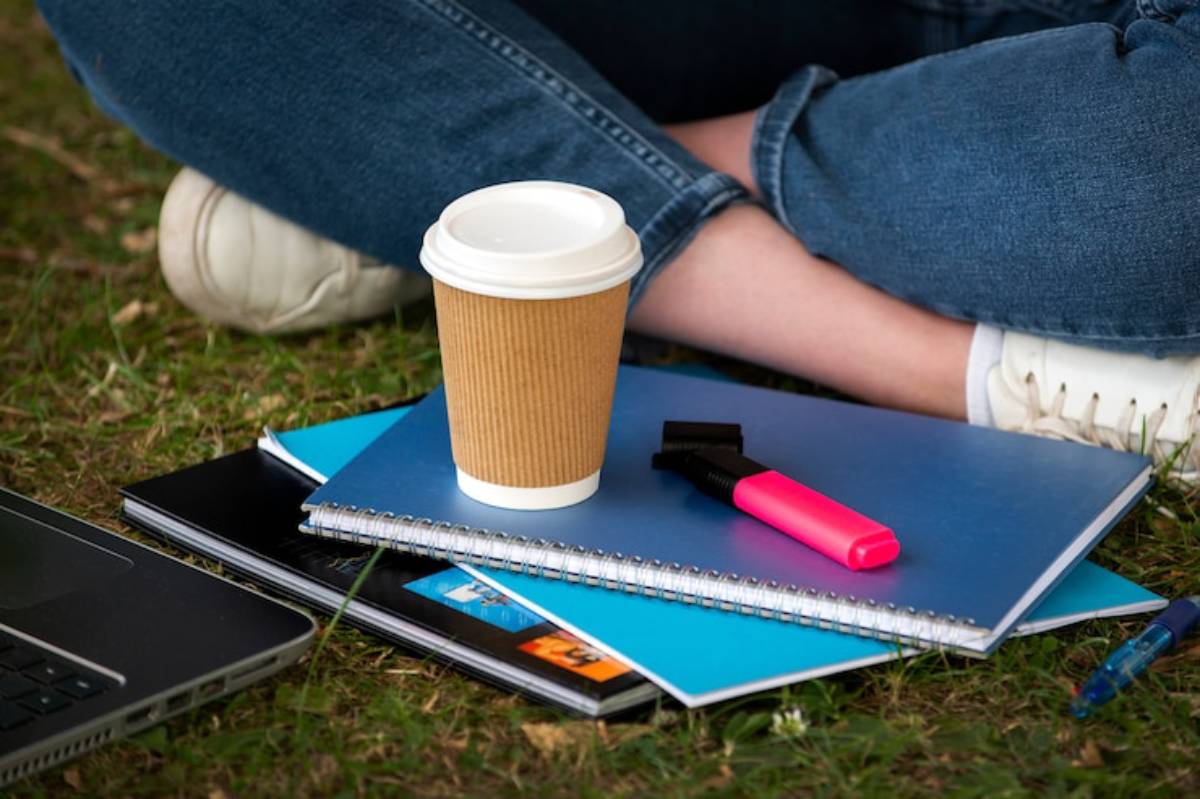 A person sits on grass with coffee, notebooks, a laptop, and a bright pink highlighter, enjoying a study session outdoors.