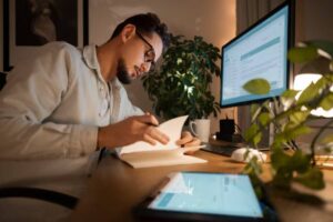 A person writes in a notebook at a desk with a computer and tablet, surrounded by a houseplant in a cozy, lit room.