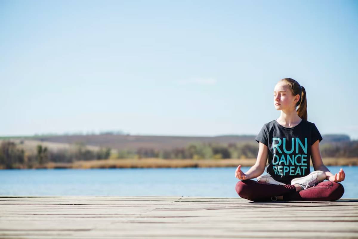 A person sits cross-legged on a wooden dock, practicing mindfulness by a calm blue lake under a clear sky.