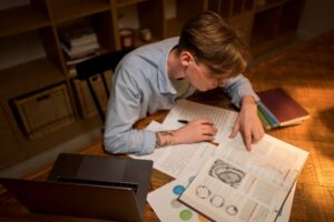 A person studies with a laptop and various books open on a wooden table, taking notes from a detailed textbook.