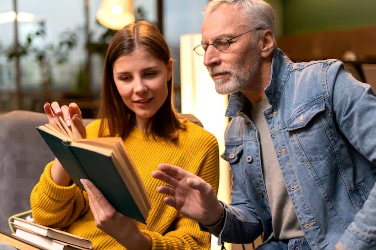 A woman in a yellow sweater reads a book while a man in a denim jacket gestures as they discuss the content in a cozy setting.
