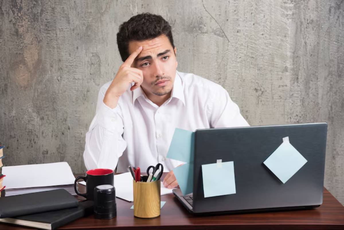 A person sits at a desk, holding a phone, surrounded by books, stationery, and a laptop with sticky notes, against a textured wall.