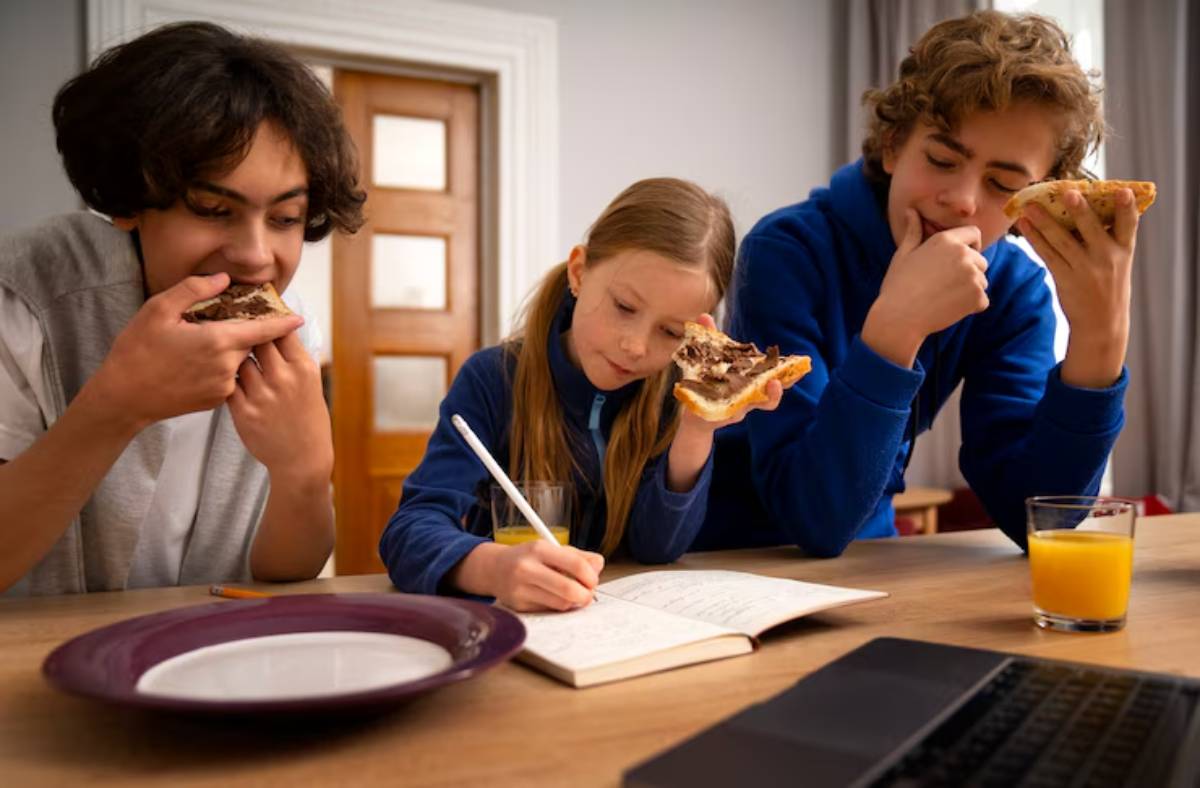Three individuals sit at a table with a laptop, eating breakfast and studying, including toast and orange juice.