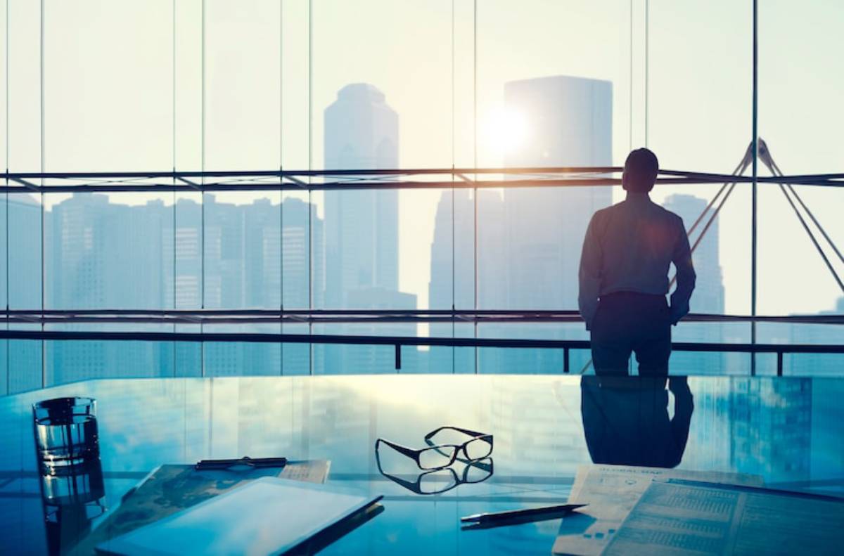 A businessman gazes out of a glass office overlooking a city skyline at sunset, with papers and glasses on a reflective desk.