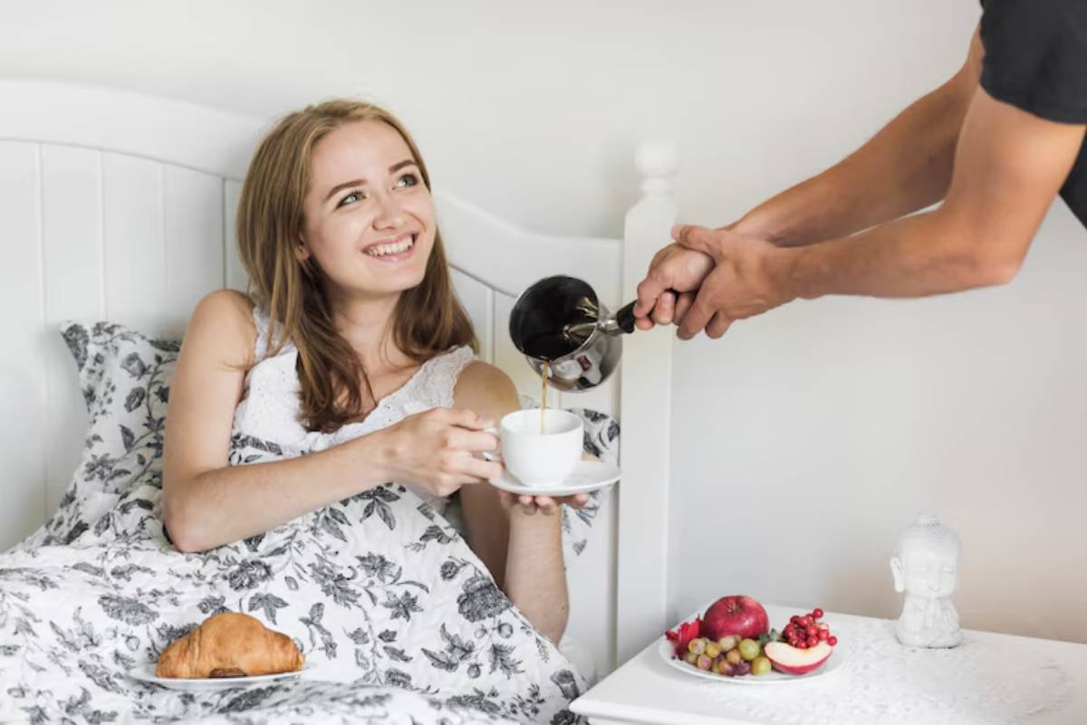A person in bed, wrapped in a floral blanket, enjoys a cozy breakfast with a croissant and fresh fruit, while coffee is being poured.