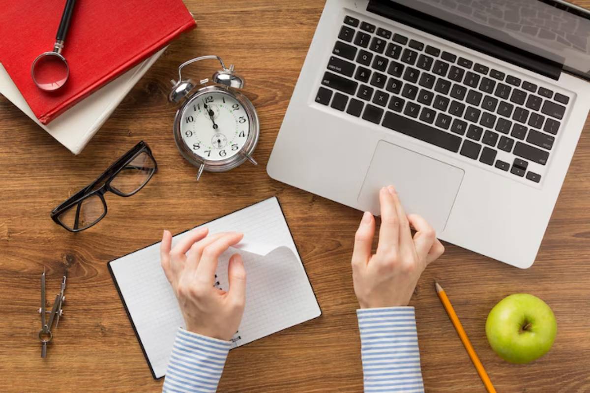 A person's hands interact with a laptop on a wooden desk, surrounded by books, an alarm clock, glasses, and an apple.