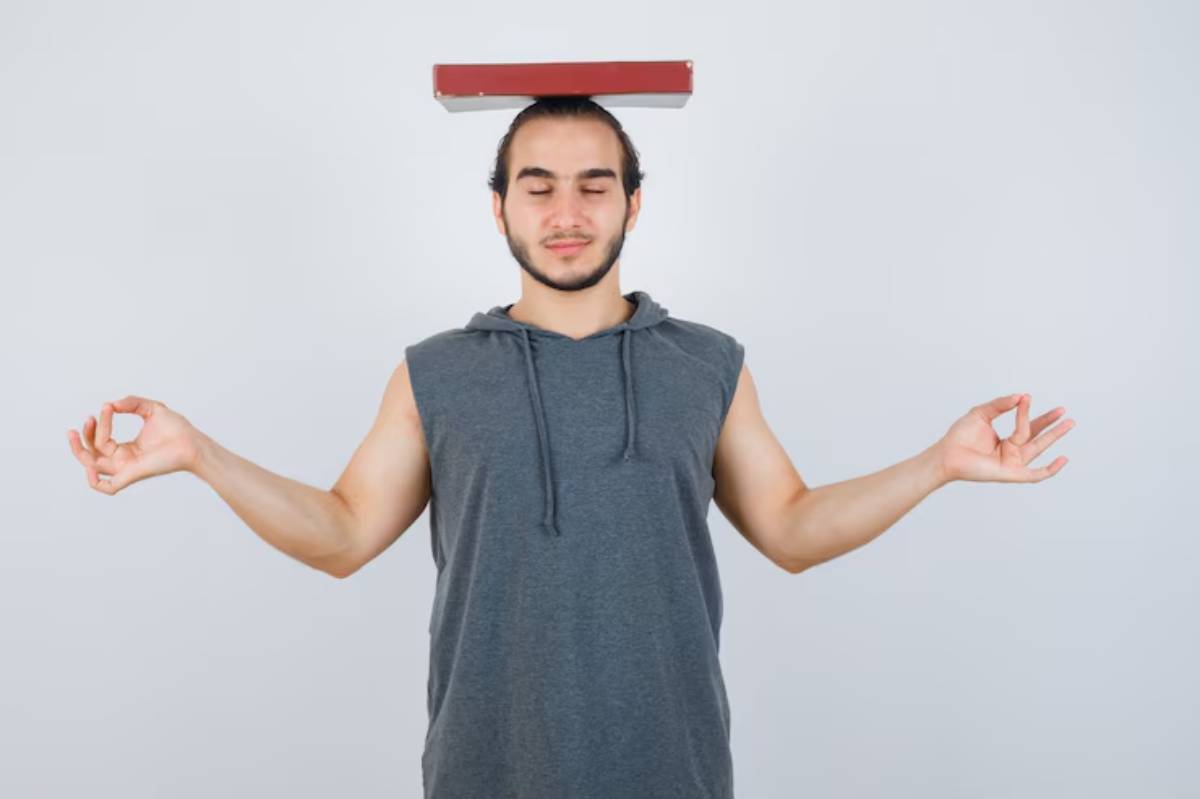 A person in a gray sleeveless hoodie balances a red book on their head, with arms outstretched in a meditative pose against a plain background.