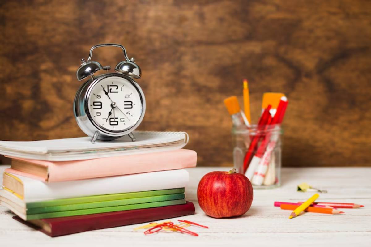 A classic alarm clock sits atop colorful stacked books, next to a shiny red apple and a jar filled with assorted writing tools.