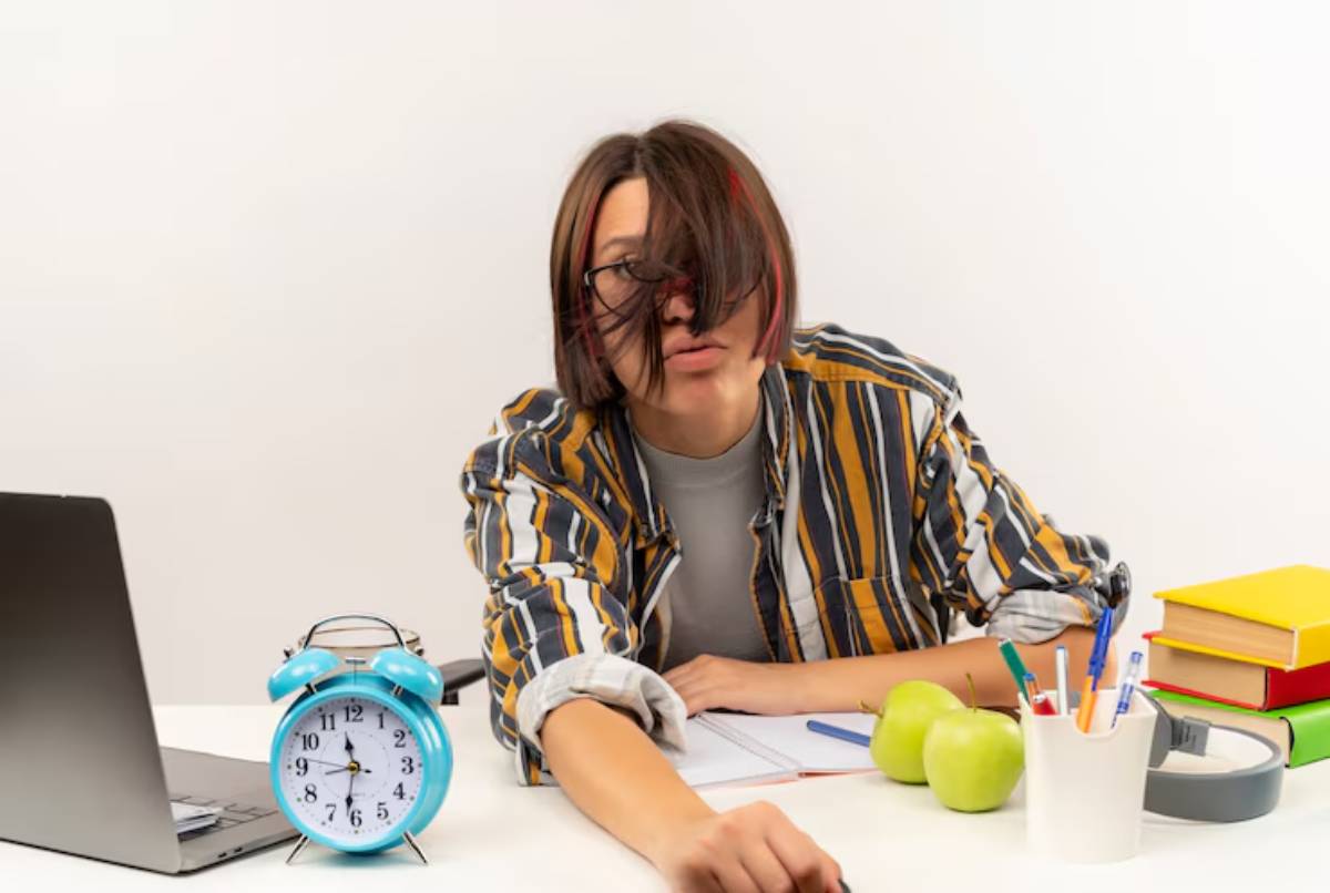 A student in a striped shirt sits at a desk with a laptop, alarm clock, apples, and colorful stationery.