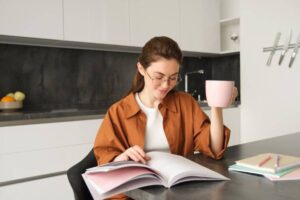 A person seated at a modern kitchen table, holding a pink mug, surrounded by books and pastel-colored stationery.