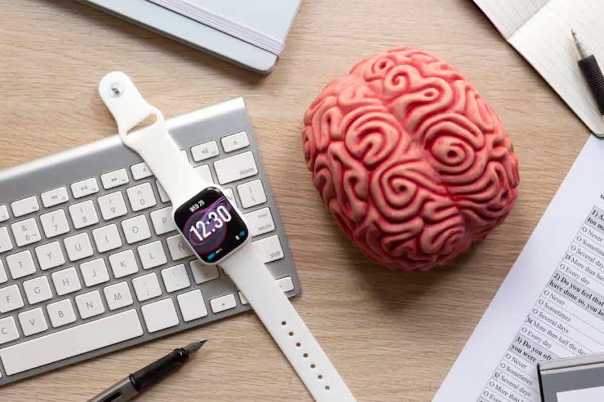 A smartwatch displays 12:30 beside a decorative brain model on a wooden desk, accompanied by a keyboard and notebook.