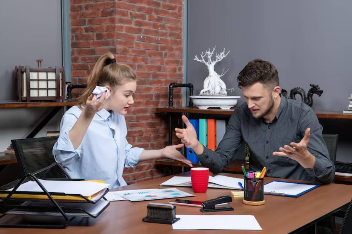 A woman and a man engaged in a heated discussion at a desk filled with papers, a red cup, and office supplies.