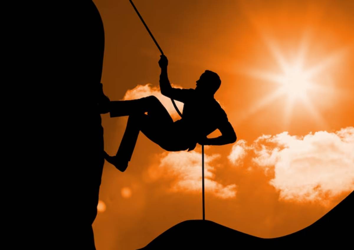 A silhouette of a climber rappelling against a vibrant orange sunset, with dramatic clouds and a bright sun.