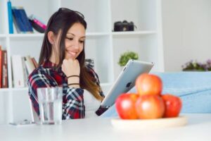 A woman relaxes on a couch, using an iPad while a bowl of red apples and a glass of water sit nearby in a bright room.