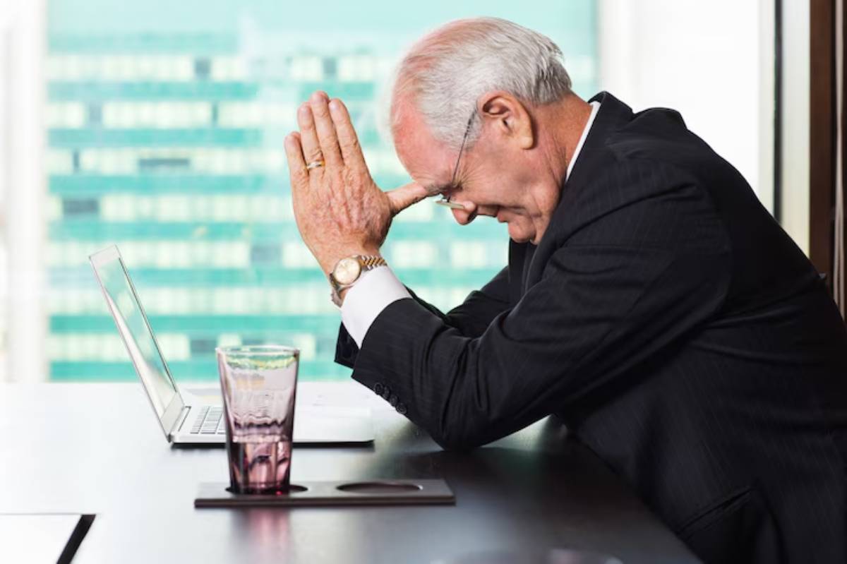 A stressed businessman in a suit rests his hands on his forehead, with a laptop and a glass of water on a conference table.