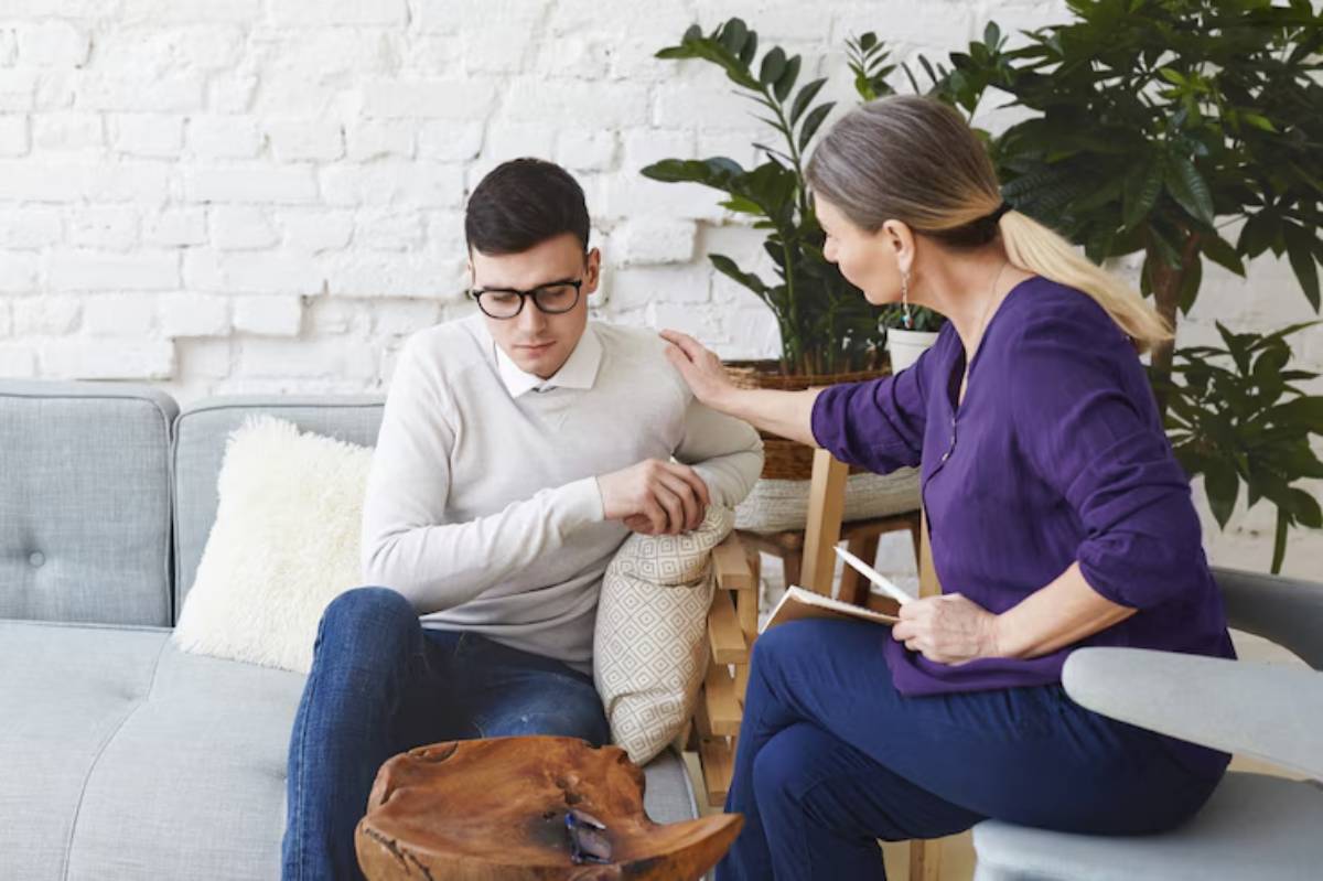 A woman in a purple blouse engages in conversation with a man on a gray couch, surrounded by greenery and a cozy interior.