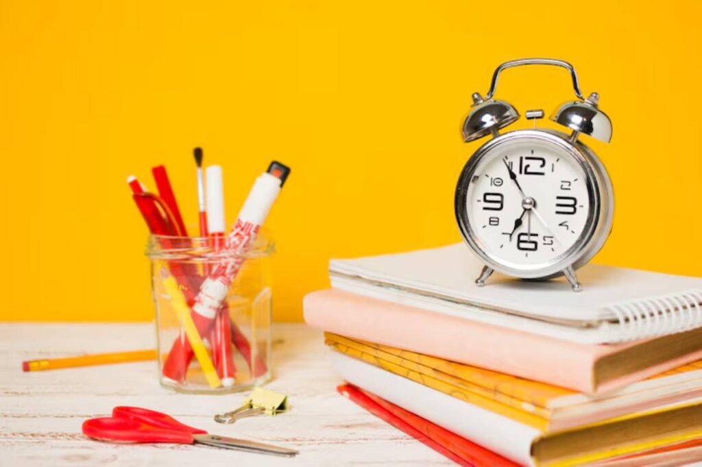 A silver alarm clock sits atop colorful notebooks, alongside a jar of red and yellow art supplies, against a bright yellow background.