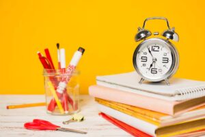 A silver alarm clock sits atop colorful notebooks, alongside a jar of red and yellow art supplies, against a bright yellow background.