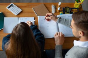 Two people studying together at a wooden table filled with notebooks, a calculator, and stationery. One writes notes, the other solves problems.