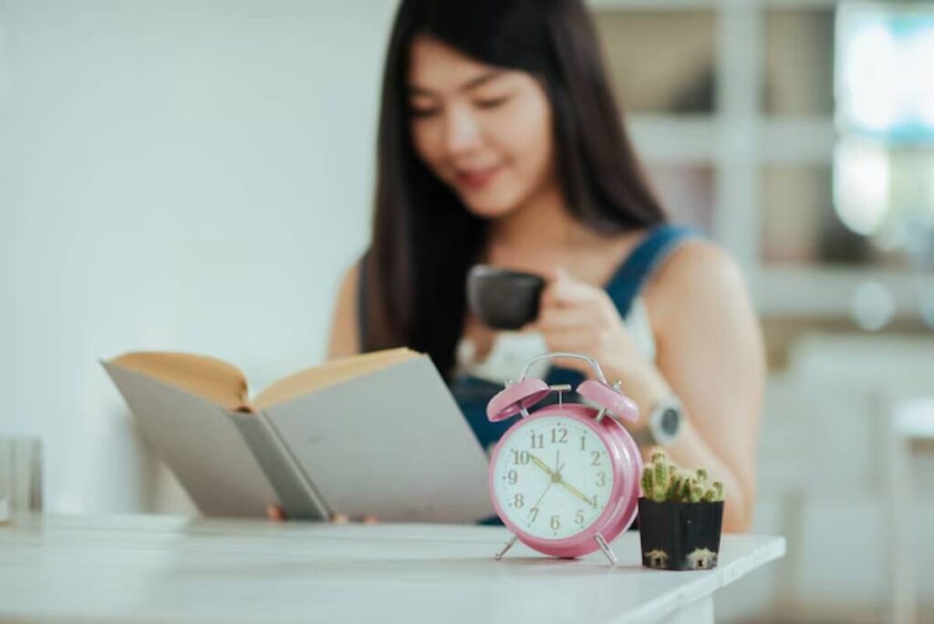 A woman holds a cup while reading a book. A pink alarm clock and a small potted cactus are on the table beside her.