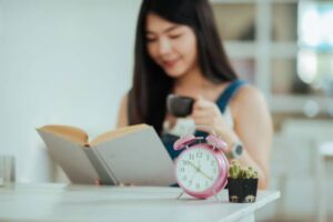A woman holds a cup while reading a book. A pink alarm clock and a small potted cactus are on the table beside her.