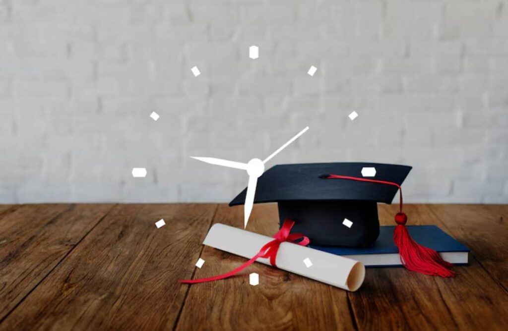 A graduation cap rests atop a book beside a rolled diploma with a red ribbon, set against a textured white wall and wooden surface.