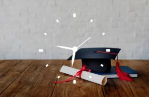 A graduation cap rests atop a book beside a rolled diploma with a red ribbon, set against a textured white wall and wooden surface.